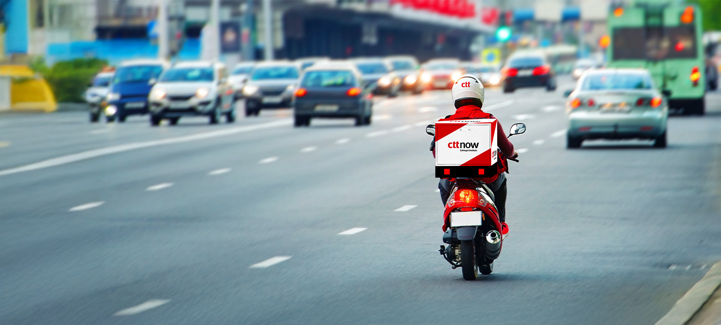 Duas pessoas numa sala, uma está sentada ao computador com livros e papéis, outra está com a farda dos CTT com uma caixa na mão.